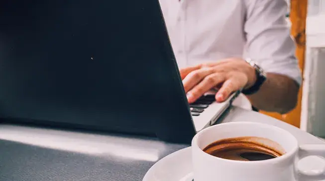 homem digita em computador com chícara de café ao lado